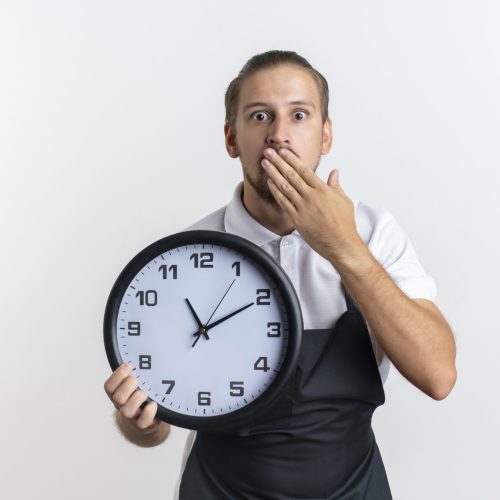 surprised young handsome barber wearing uniform holding clock and putting hand on mouth isolated on white background with copy space